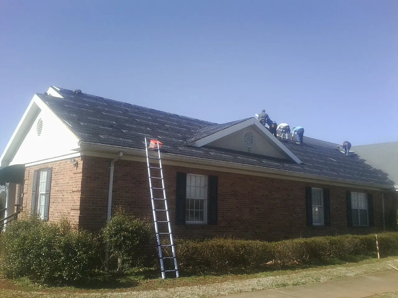 Crew tearing shingles off a brick ranch roof with a ladder set — Emergency Roof Repair work by our crew in Dacono