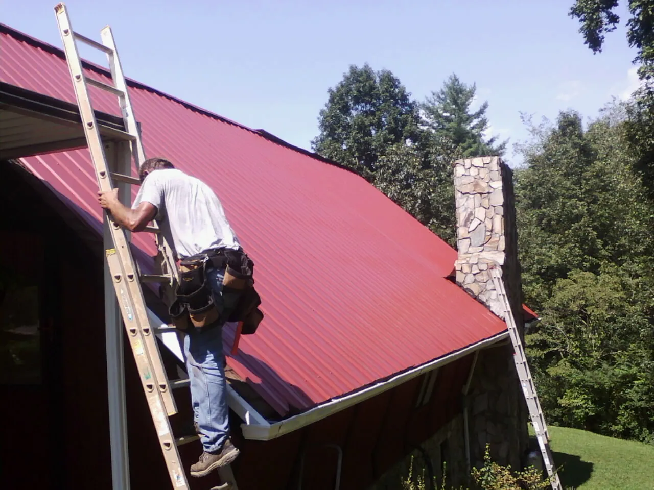 Roofer on a ladder beside a red metal roof and stone chimney — Soffit & Fascia Repair by Roofing Brothers in Dacono